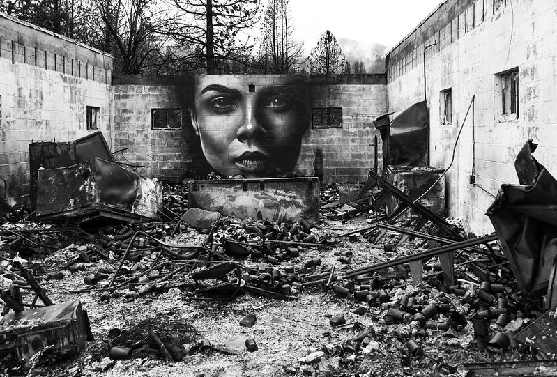 black and white image of a mural showing a woman's face painted on a wall of a burned out building with no roof. Blackened, burned cans and furniture lie in the foreground