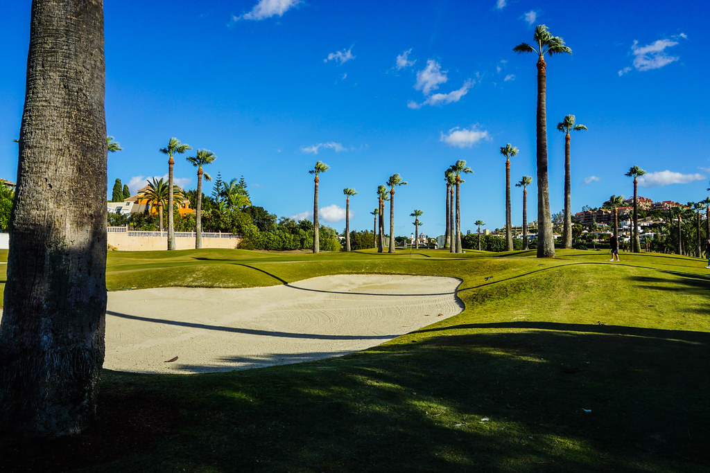 A grass portion of a golf course with a big patch of sand in the middle. Palm trees are spread out all over the grass. On the left side, a stone wall, partially covered in vegetation, and a villa showing from behind it. On the right side, a man wearing a black t-shirt and blue shorts is walking on the grass. On the far right side, buildings are visible from behind a few trees.