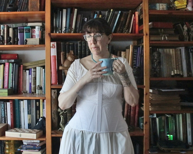Femme portant un corset blanc type 1810 environ et tenant une tasse de thé, devant une bibliothèque
