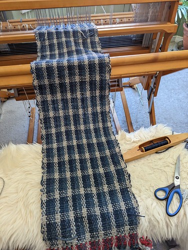 Length of cloth in loom state is laid across irieknit's loom bench and is a 3-colour plain weave check with undyed handspun Shetland wool and 2 shades of natural indigo.  A boat shuttle with the dark shade of indigo is on the loom bench and so is a pair of scissors.