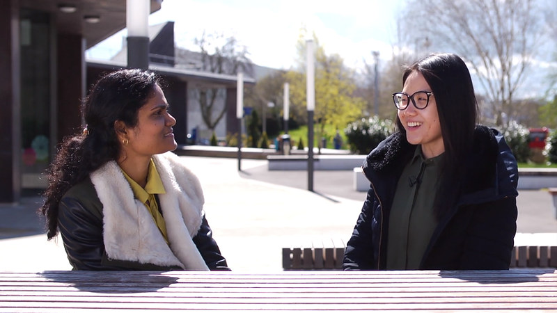 Two female Malaysian students talk to one another, seated outside at a table. 