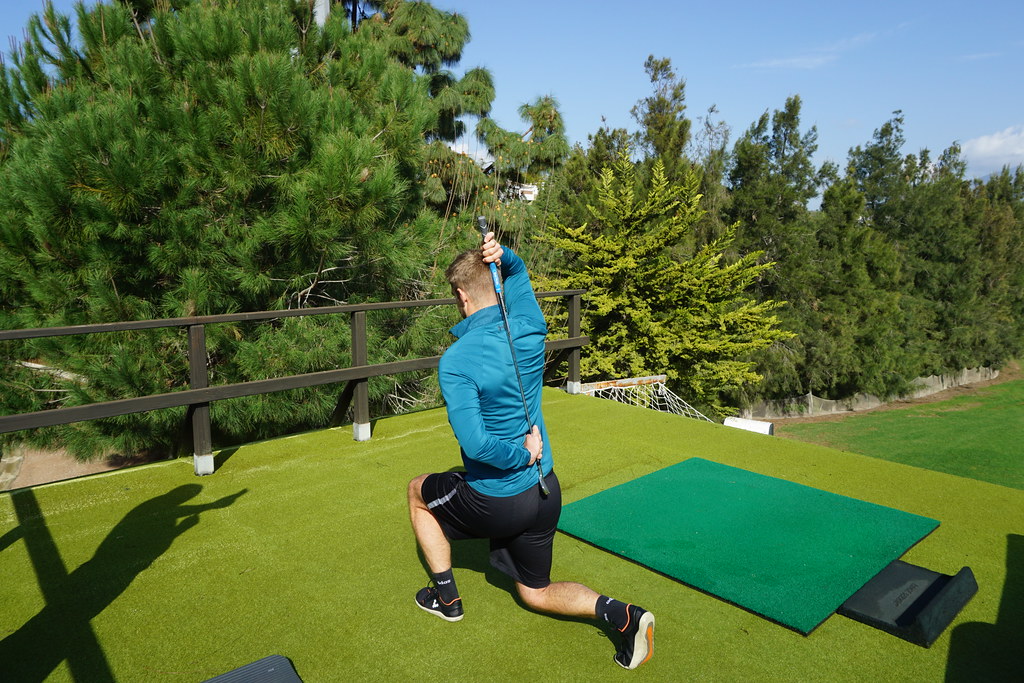 A man, on a golf driving range, wearing a blue sweatshirt, black shorts and black trainers is lunging, left foot forward. He is holding a golf club against his back with both his hands. Next to him, on the right, there’s an exercise mat. Green grass is visible on the right of the driving range. A tree line is in the far background.