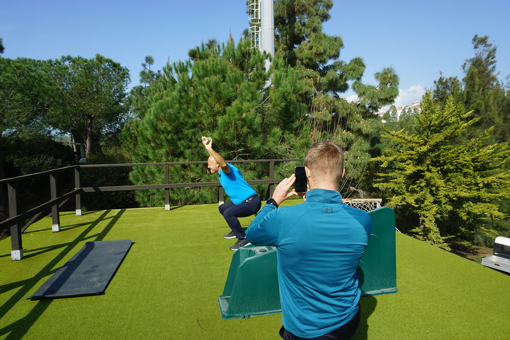 2. Two men. The one in the background is wearing a light blue t-shirt, dark blue trousers and trainers, while doing squats. The man in the foreground is wearing a blue sweatshirt, standing with his back against the camera, taking a photo of the other man with his phone. They are on a green golf driving range. Trees are forming the background of the photo.