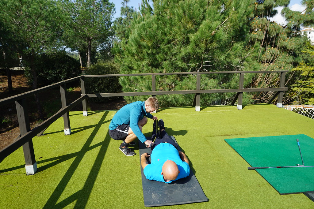 Two men on a golf course driving range. One lying down on a mat, wearing a baby blue t-shirt, navy blue trousers and trainers. The other man, wearing a blue sweatshirt, black shorts and trainers, is squatting next to him, with a measuring tape. On a mat next to them, two golf clubs. The background is made of the driving range’s fence and further in the back, a line of trees.