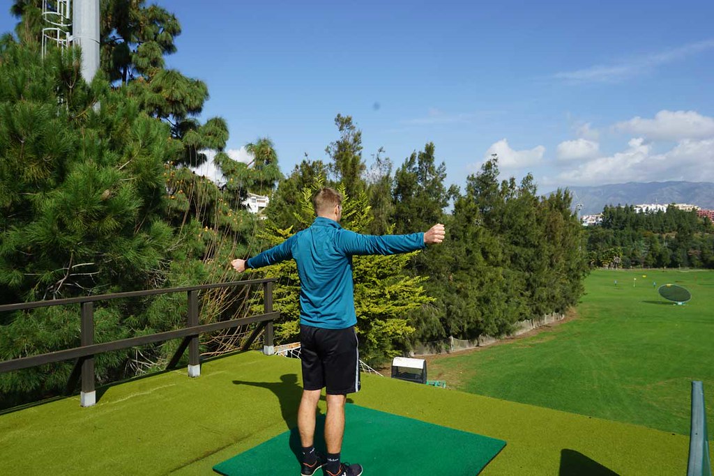 A man, standing straight on a golf driving range, with his back at the camera, arms stretched wide, looking into the distance. He’s wearing a blue sweatshirt, black shorts and trainers. On the right side of the background a golf course. On the left side and far back of the photo, trees and bushes form a fence.