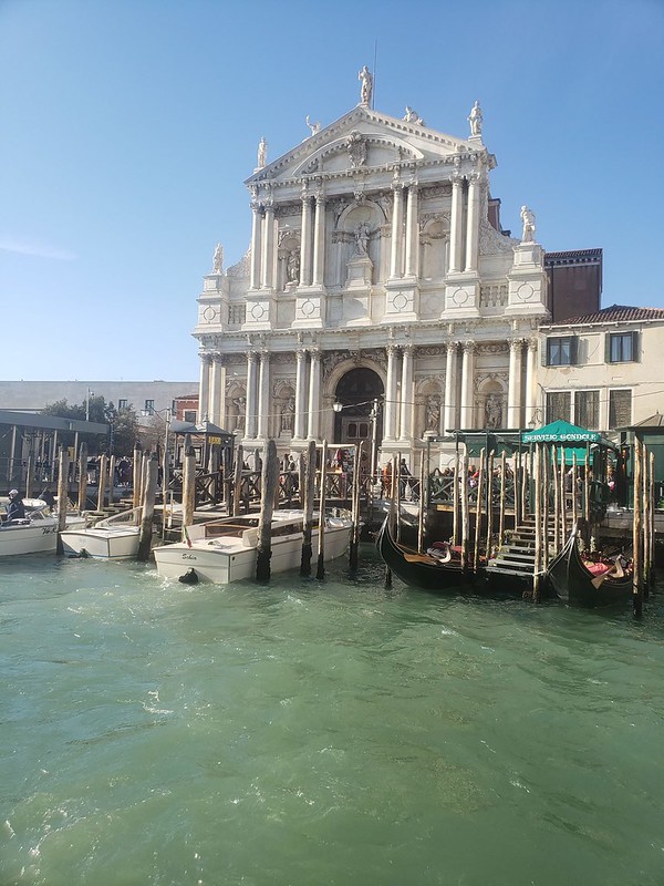 Gondolas docked on the light green water in front of a white and gold decadent building with columns and statues. 