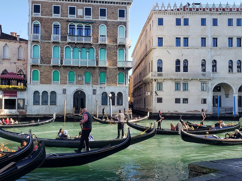 Gondoliers driving the gondolas with their paddle sticks as they transport people down a wide canal. 