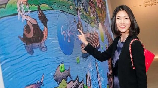 Siyang stands next to a mural map on a wall showing a river, fish and boats. She smiles towards camera.