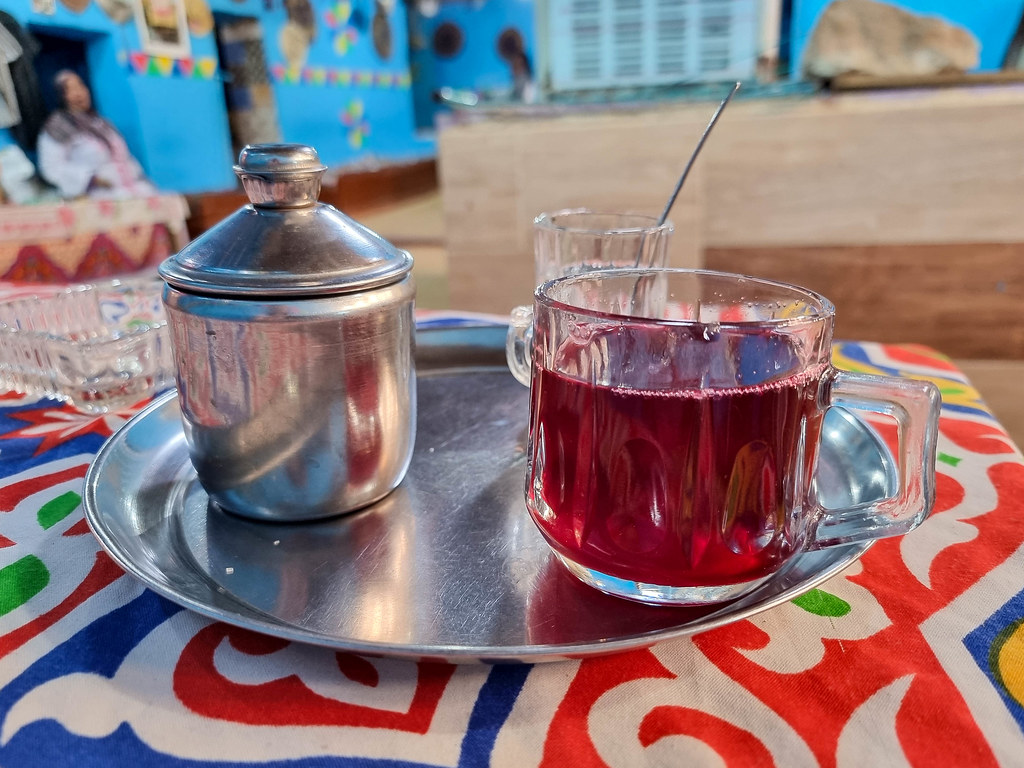 A metal tray with a cup of cold hibiscus tea, which is bright red. On the tray there is also a metal container filled with sugar and a glass with a teaspoon in it.