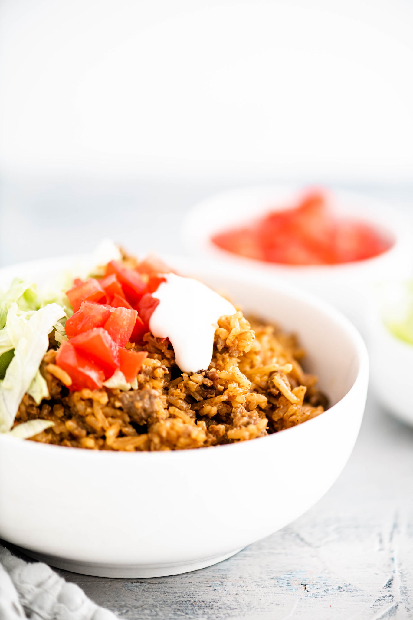 Queso rice with ground beef in a white bowl topped with shredded lettuce, tomatoes and sour cream.