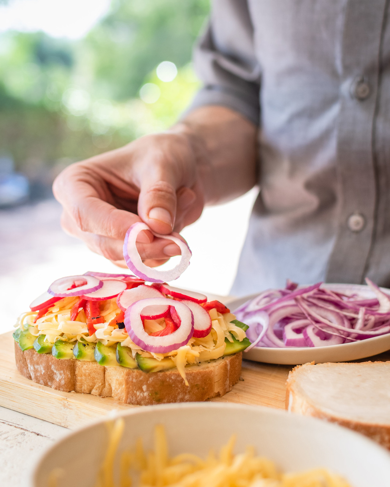 placing sliced red onions on a loaded sandwich with avocado, cheese, peppers, and radishes