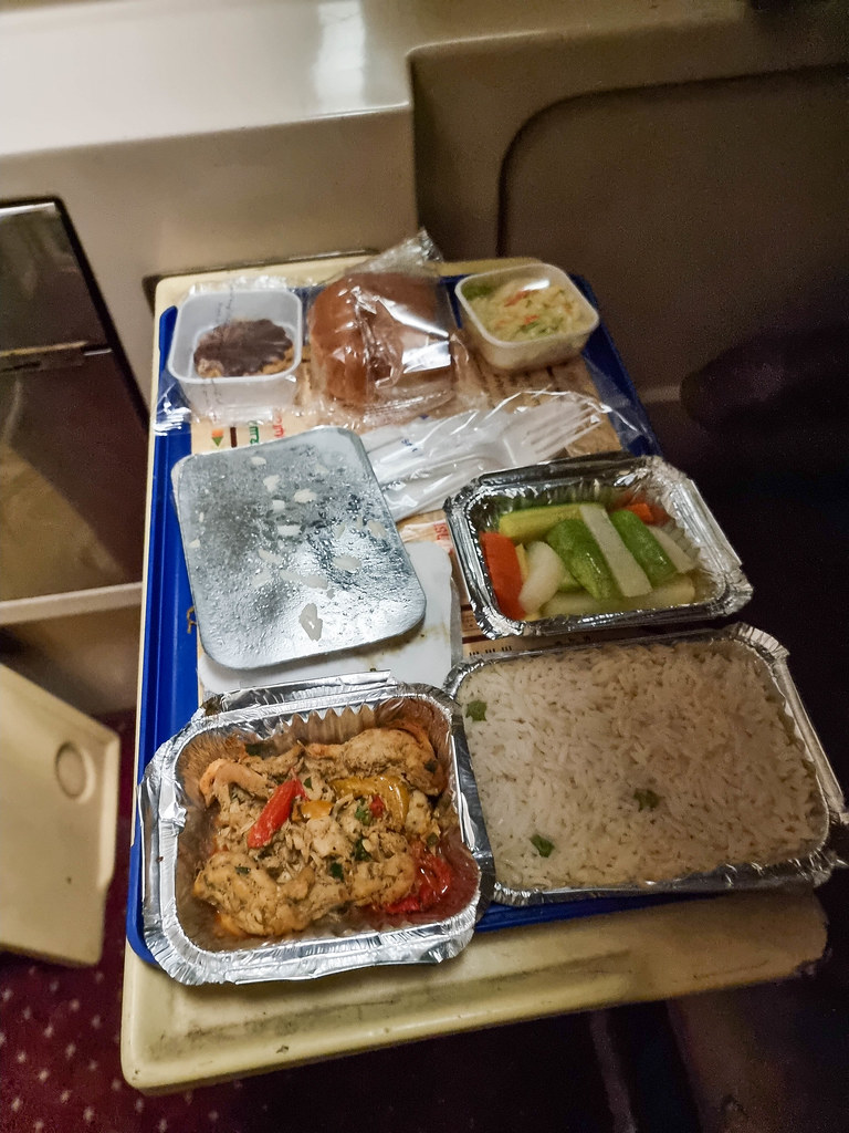 A close-up of the dinner spread on a blue tray. There is a container with chicken stew, one with a salad, one with boiled vegetables, one with rice, a bread roll and a biscuit covered in chocolate