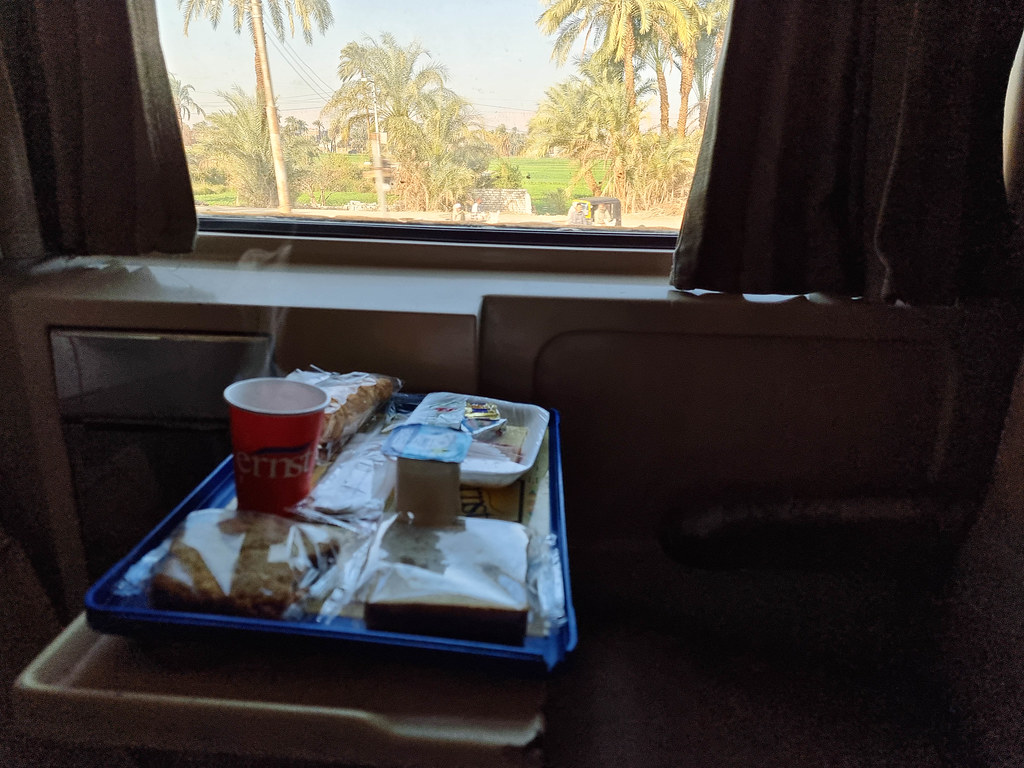 Breakfast on the train: a blue tray with a continental selection and a red cup of coffee. On the train's window you can see green palm trees and a blue sky