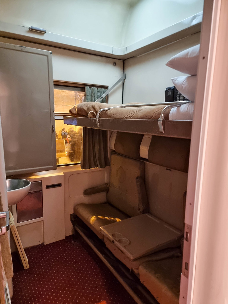 A vertical imagine of the entire cabin. The top bed is made, whilst underneath there are two chairs. In between the chairs there is a white table with spaces for two glasses in it