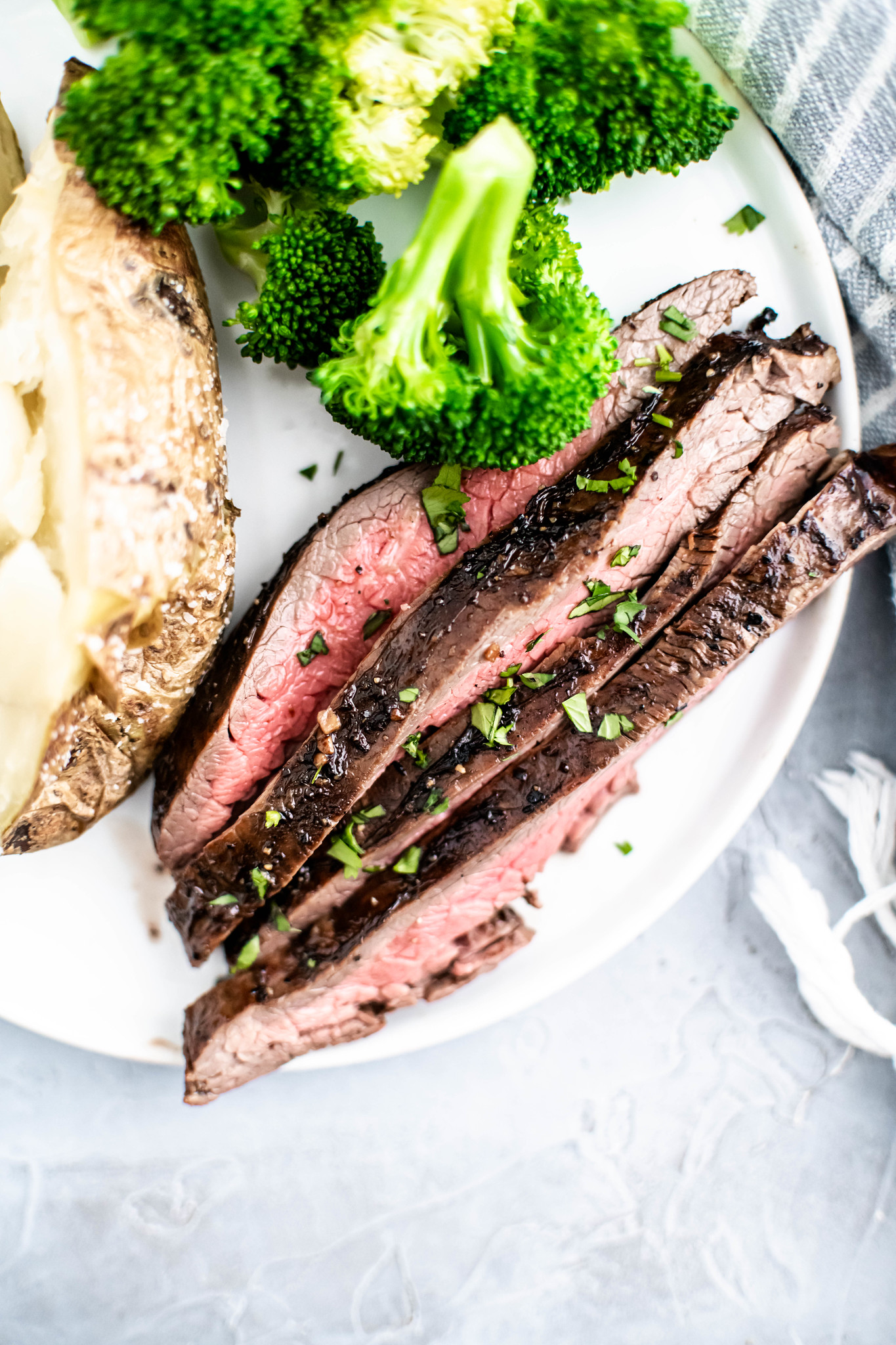Thinly sliced flank steak on a plate with a baked potato and steamed broccoli.