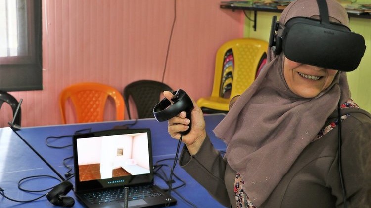 A female faces camera, smiling and wears a virtual reality headset. The laptop on a table by her shows a room.