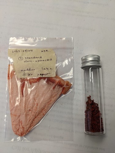 On the left lies a plastic bag labeled 3 Standard Alum Extracted Madder Lake Filter Paper containing emptied filter paper, on the right lies a clear vial filled with reddish pigment