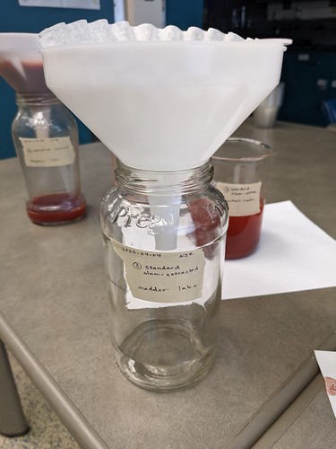 Funnel containing filter is positioned over an empty glass jar labeled Standard Alum Extracted Madder Lake
