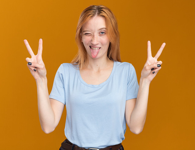 joyful young redhead ginger girl with freckles stucks out tongue and gestures victory sign isolated on orange background with copy space
