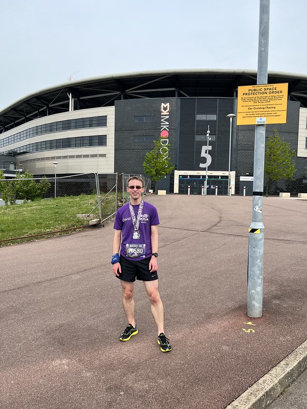 A caucasian male weating a purple t-shirt and black shorts in front of a stadium. He had a medal around his neck