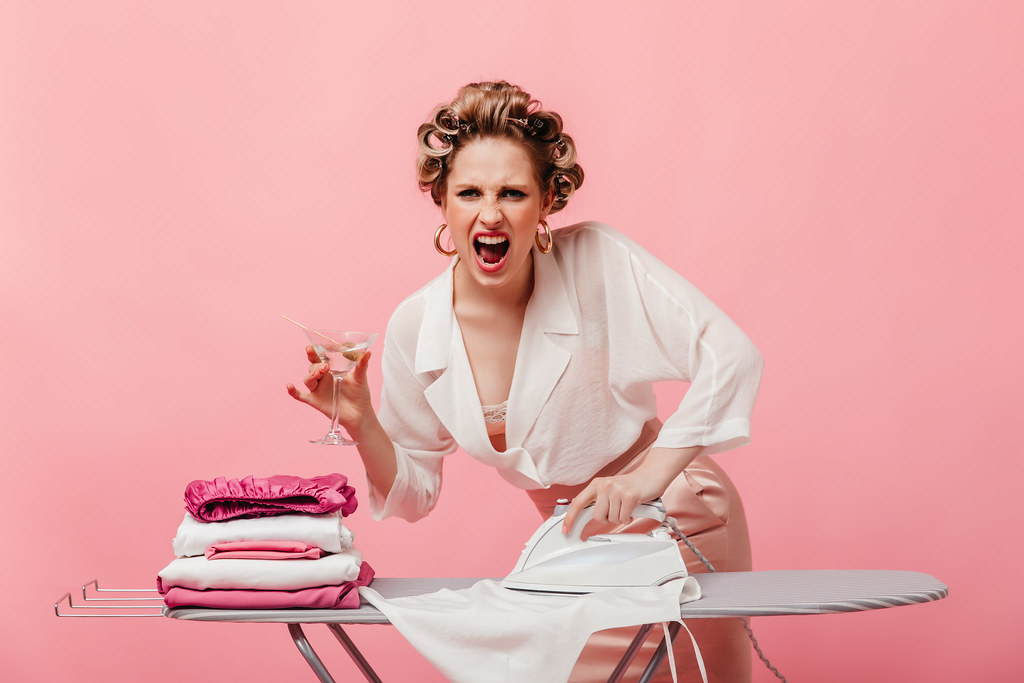 Aggressive housewife with hair curlers holds martini glass and irons white blouse.