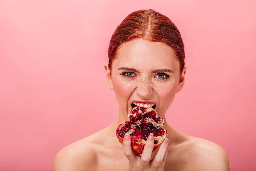 Front view of hungry woman eating pomegranate. Studio shot of naked ginger girl with garnet isolated on pink background
