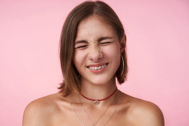 Portrait of plesant looking young short haired lady with natural makeup frowning her face while smiling cheerfully with closed eyes, isolated over pink background