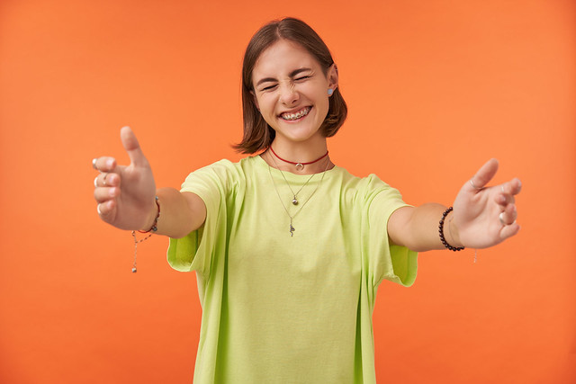 Young pretty woman squints smiling and holding her hand out for a hug. Student happy to see her friends. Wearing green t-shirt, teeth braces, bracelets, necklace. Portrait over orange back ground