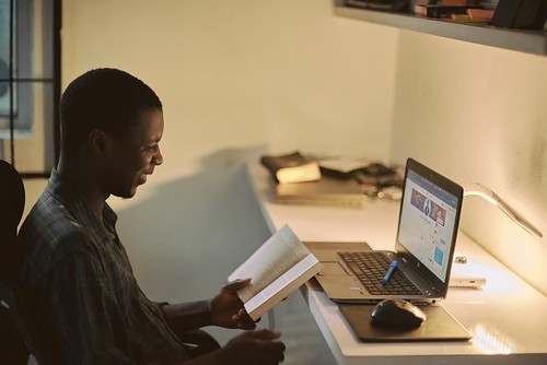 A young black man sits at his desk. A laptop computer sits in front of him. He is smiling and reading a book.