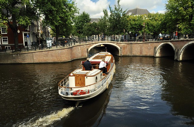 Steel ship with beautiful wooden trim and accents with round porthole windows in the lower living part of the ship it could be used as a small recreational or tour ship in this canal with bridges across it & steel railings with lots of bicycles against it