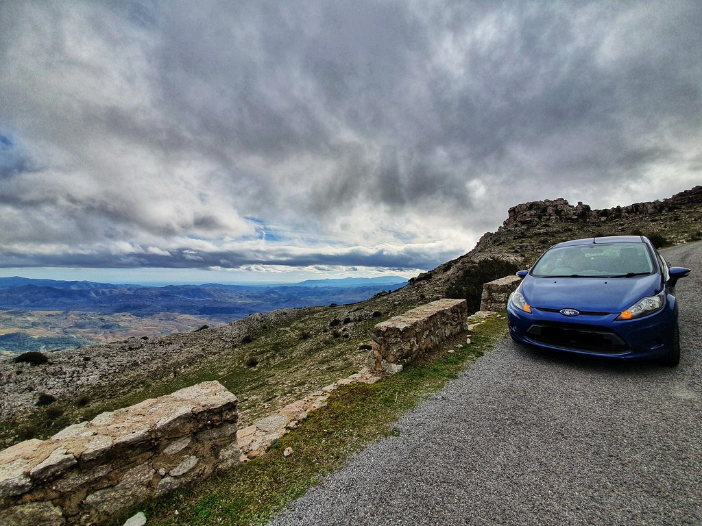 A blue car parked on the side of a road, on a mountain. On the left hand side there is a beautiful view on the valleys below. The sky is clouded.