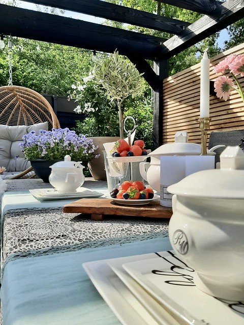 Gedekte tuintafel houten serveerplank Riviera Maison servies zomerfruit op etagere hangstoel aan zwarte veranda