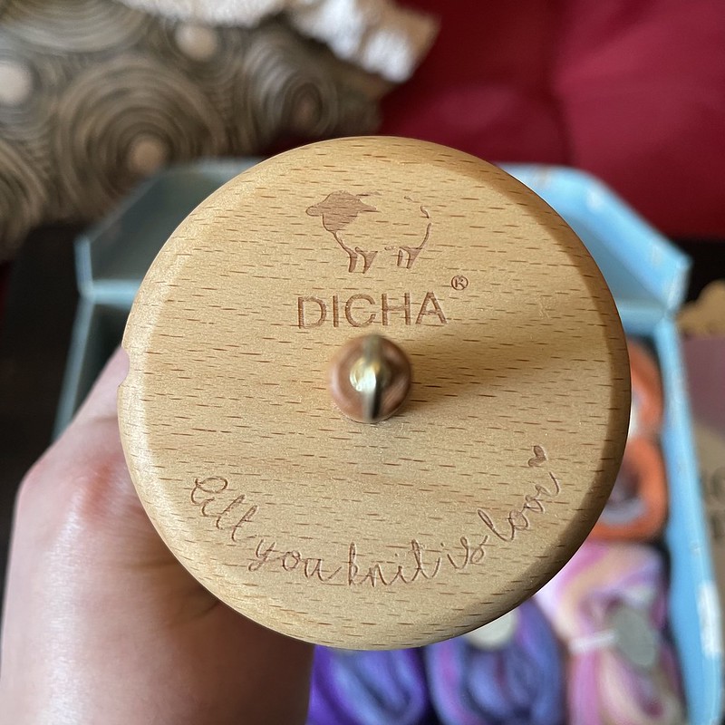 the top of a wooden drop spindle with the image of a sheep, "Dicha" and ""All you knit is love" engraved