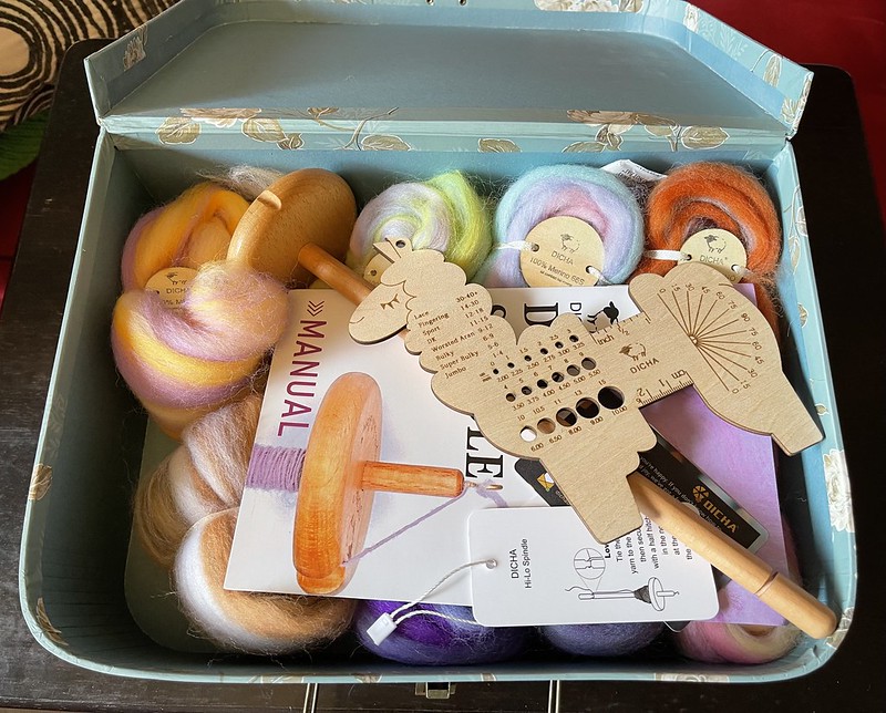 an open box with a llama-shaped yarn gauge, a drop spindle, a manual, and several bundles of colorful wool
