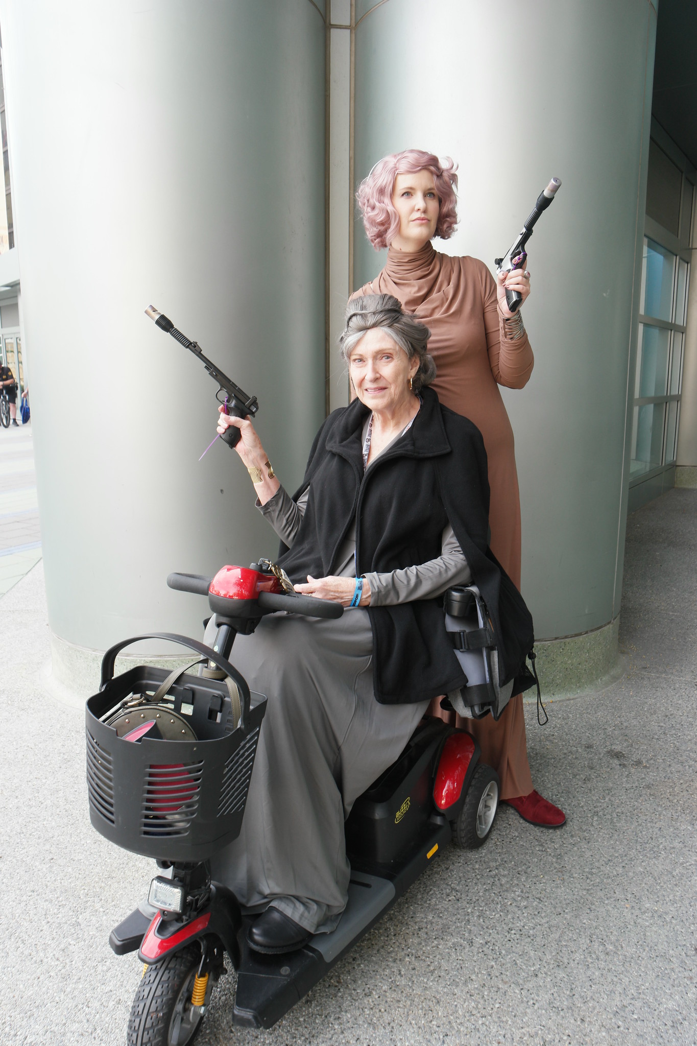 An older white woman with grey hair sits on a scooter dressed as General Leia. Behind her stands another woman with pink hair. They are each holding up blaster pistols.
