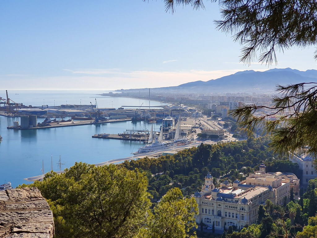 The panorama from Gibralfaro viewpoint, over the city, the sea on the left, and the mountains in front, in the far. There is a hazy mist raising over the city, near the mountains
