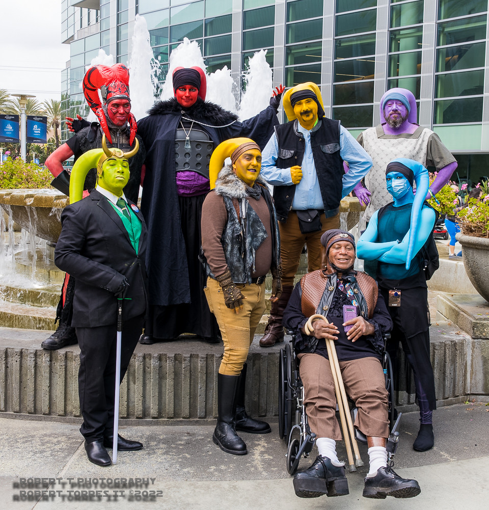 A group of Star Wars cosplayers are dressed up as Twi'Lek aliens and each have skin painted different colors with matching "head-tail" tentacles. One cosplayer is green, is wearing a suit, and has a Loki-style horned helmet and is holding a cane. Another with naturally dark skin and match head-tails is seated in a wheelchair.
