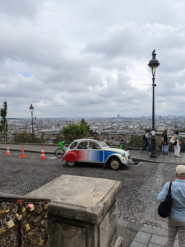 Deux chevaux car painted in the colours of the French flag driving past the view of Paris from the Montmartre.
