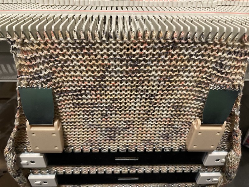 a body panel of an orangey machine knit sweater on a flat bed knitting machine with weights hanging from the fabric