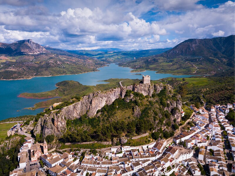 An aerial view of zahara de la sierra, with the castle on top of the hill, and the reservoir in the far end
