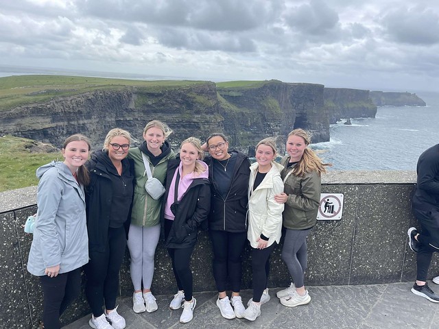 Seven students pose in front of a stone wall with the Cliffs of Moher behind them.