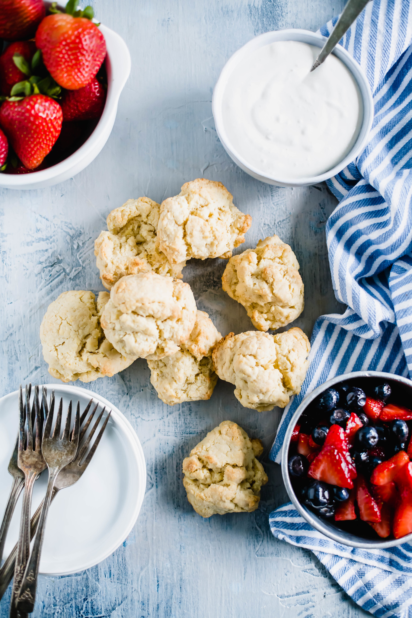 Shortcake biscuits on a blue grey background with a bowl of macerated strawberries and blueberries on the right and a bowl of whipped cream.