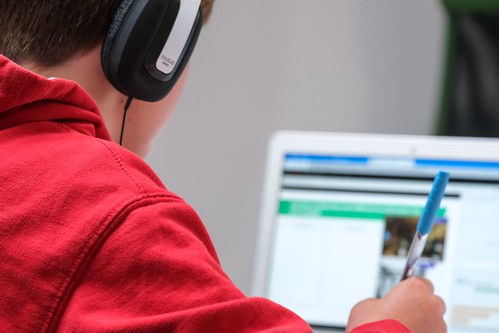A person wearing over-ear headphones in a red sweatshirt holds a pen. They look at the computer screen in front of them - How to Study for the SAT: Free Resources, Advice, & More