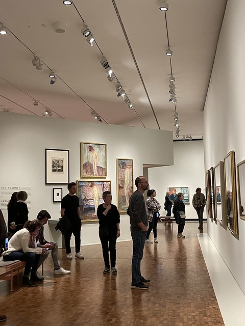 People look at framed artwork on the white walls of an art gallery.