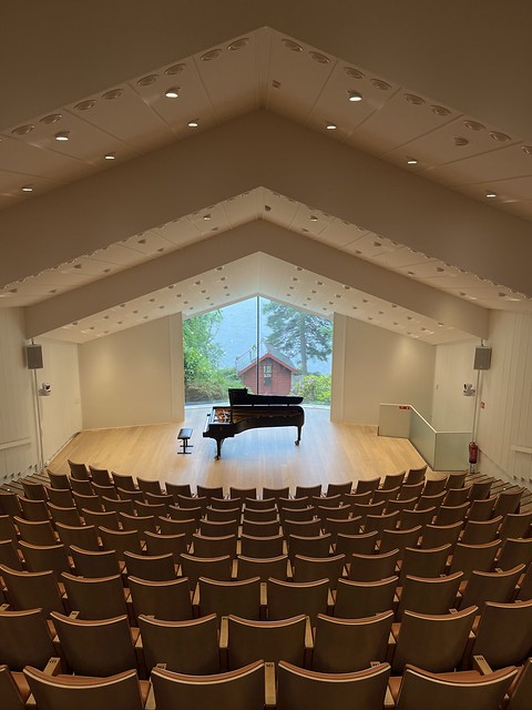 Rows of chair slope down to a stage with a single piano. A small red hut is visible through large class windows beyond the stage.