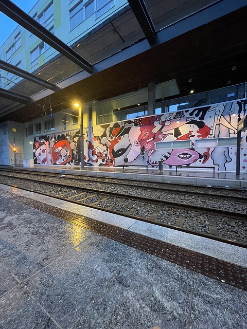 A large mural is painted on the wall behind the tracks at a tram stop.