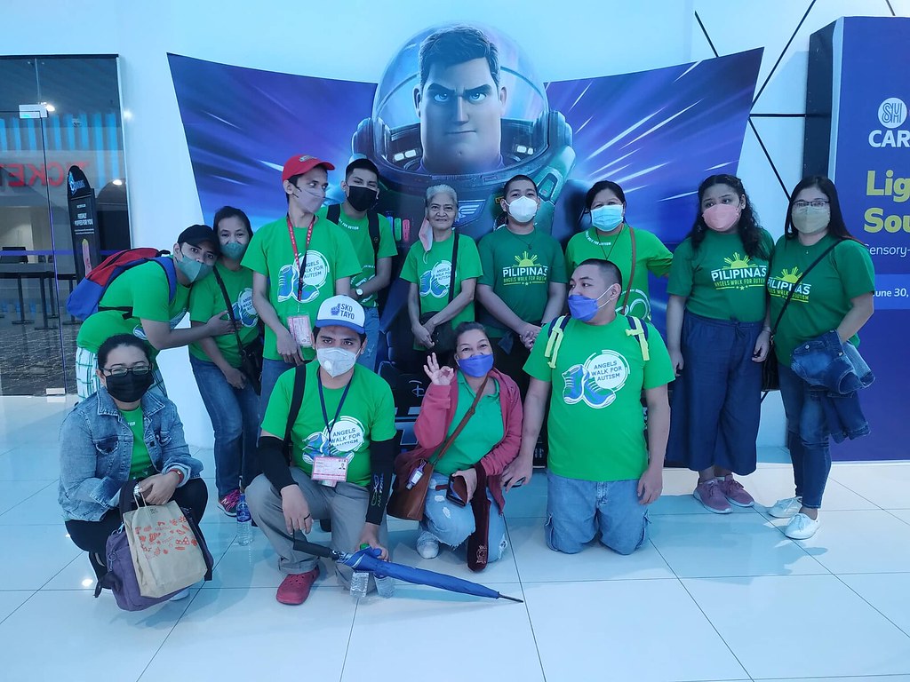 ASP Family mostly wearing 2021 Angels Walk T-Shirts, three wearing 2019 and 2020 Angels Walk T-Shirts and one wearing maong jacket standing in front of Lightyear banner for group photo.