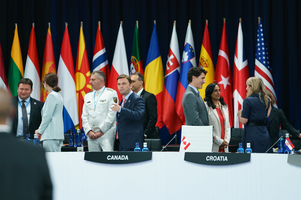 Dignitaries stand in small groups behind a large table, country flags in the background