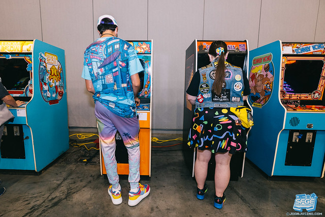 Photo of two adults in colorful, 80s themed clothing playing vintage and retro arcade games.