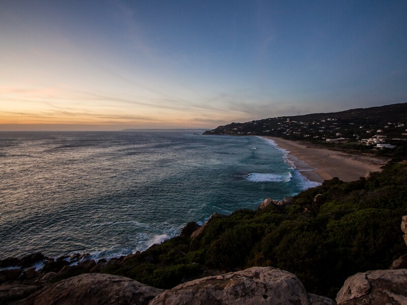 A photo of the beach taken just after sunset, from the cliffs above it. There is nobody on the beach. The beach is surrounded by thick green vegetation.